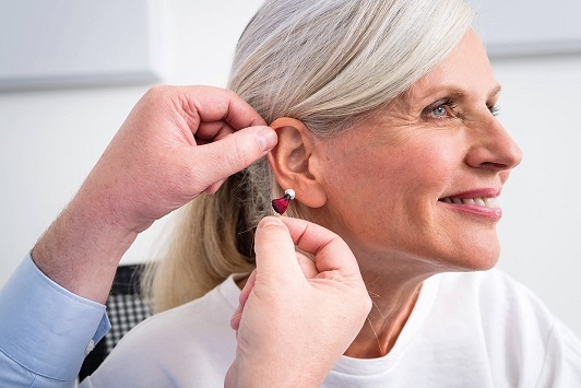 Ältere Frau bekommt ein modernes Hörgerät ins Ohr eingesetzt, Hörhilfe Akustiker Praxis.